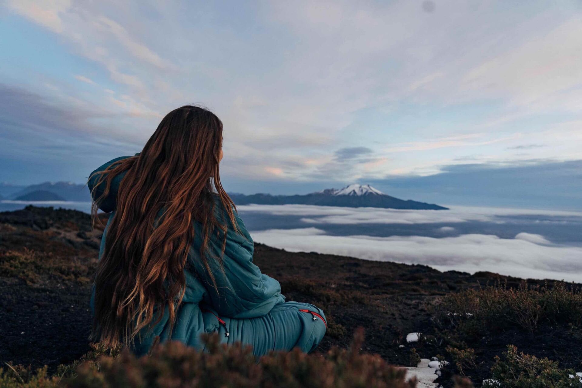 Junge Frau sitzt in ihrem warmen, tragbaren Schlafsack auf einem Gipfel und geniesst die Aussicht auf die wolkenverhangenen Berggipfel in der Ferne