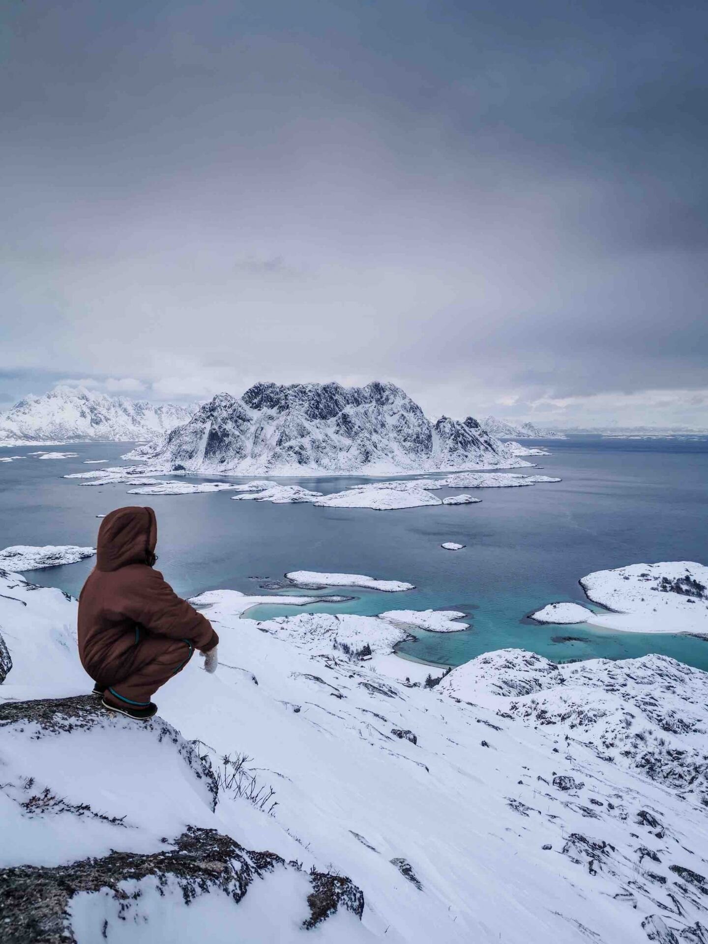 vor einer weiten verschneiten Meerlandschaft sitzt jemand eingepackt in seinen tragbaren Schlafsack und geniesst den Ausblick