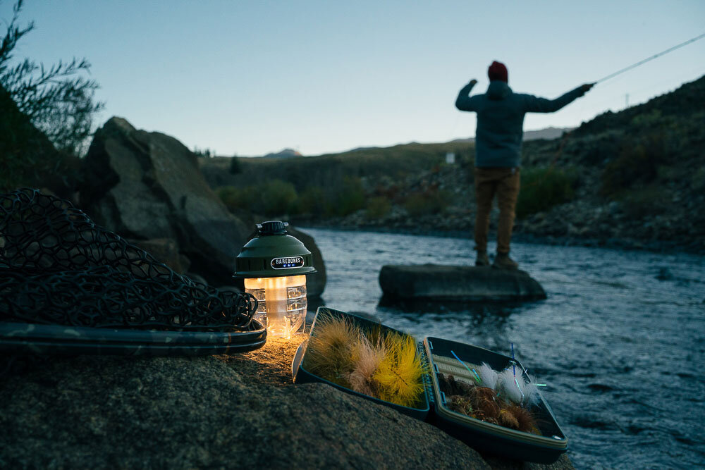 kleine wiederaufladbare und stufenlos dimmbare Campinglampe steht in der Dämmerung auf felsigem Grund und beleuchtet ein Fliegenfischerset